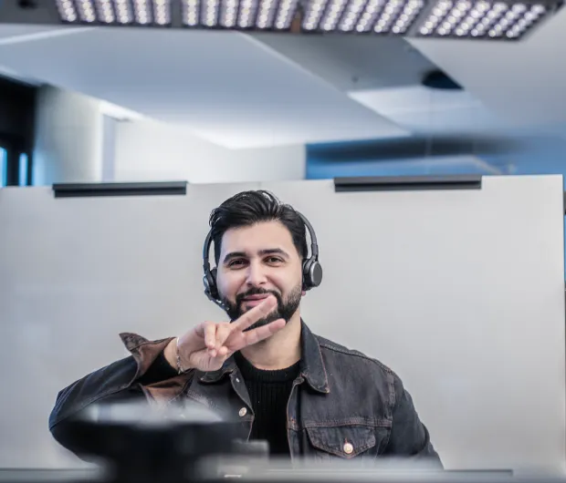 Mann mit Kopfhörer in Büro zeigt mit der Hand das Peace-Zeichen und lächelt freundlich.