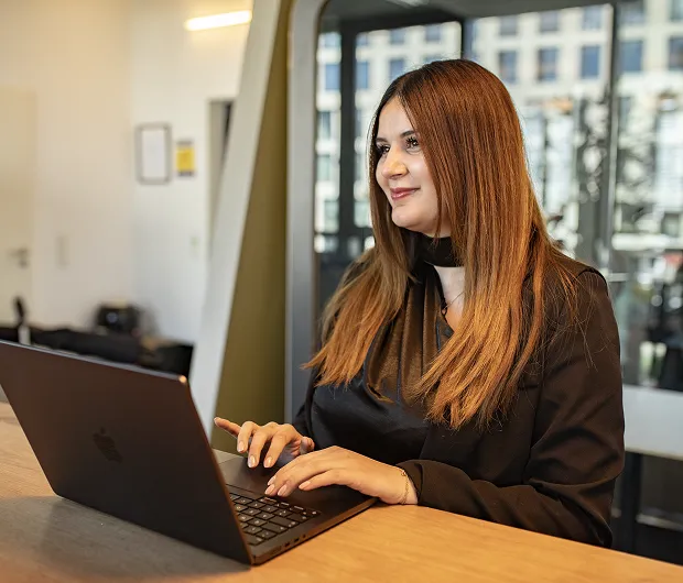 Frau mit braunen Haaren lächelt und arbeitet an einem Laptop an einem Holztisch im Büro.