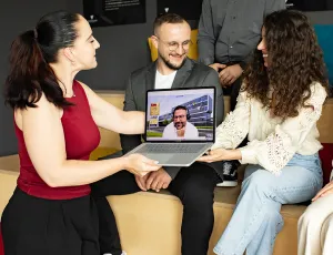 Drei Personen lachen und zeigen auf einem Laptop eine virtuelle Person während eines Videocalls.
