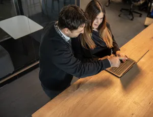 Zwei Kollegen stehen an einem Schreibtisch, einer zeigt auf die Tastatur, während sie über die Arbeit sprechen.