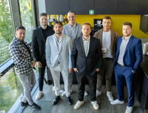 Sieben Männer in Business- und Freizeitkleidung posieren in einem modernen Büro vor einer gelben Wand.