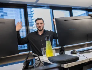 Mann steht hinter zwei großen Monitoren und hebt die Hände, moderne Büroumgebung mit Glasfenstern.