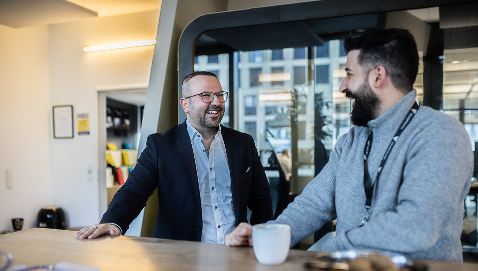 Zwei Männer lachen und unterhalten sich an einem Tisch in einem modernen Büro.