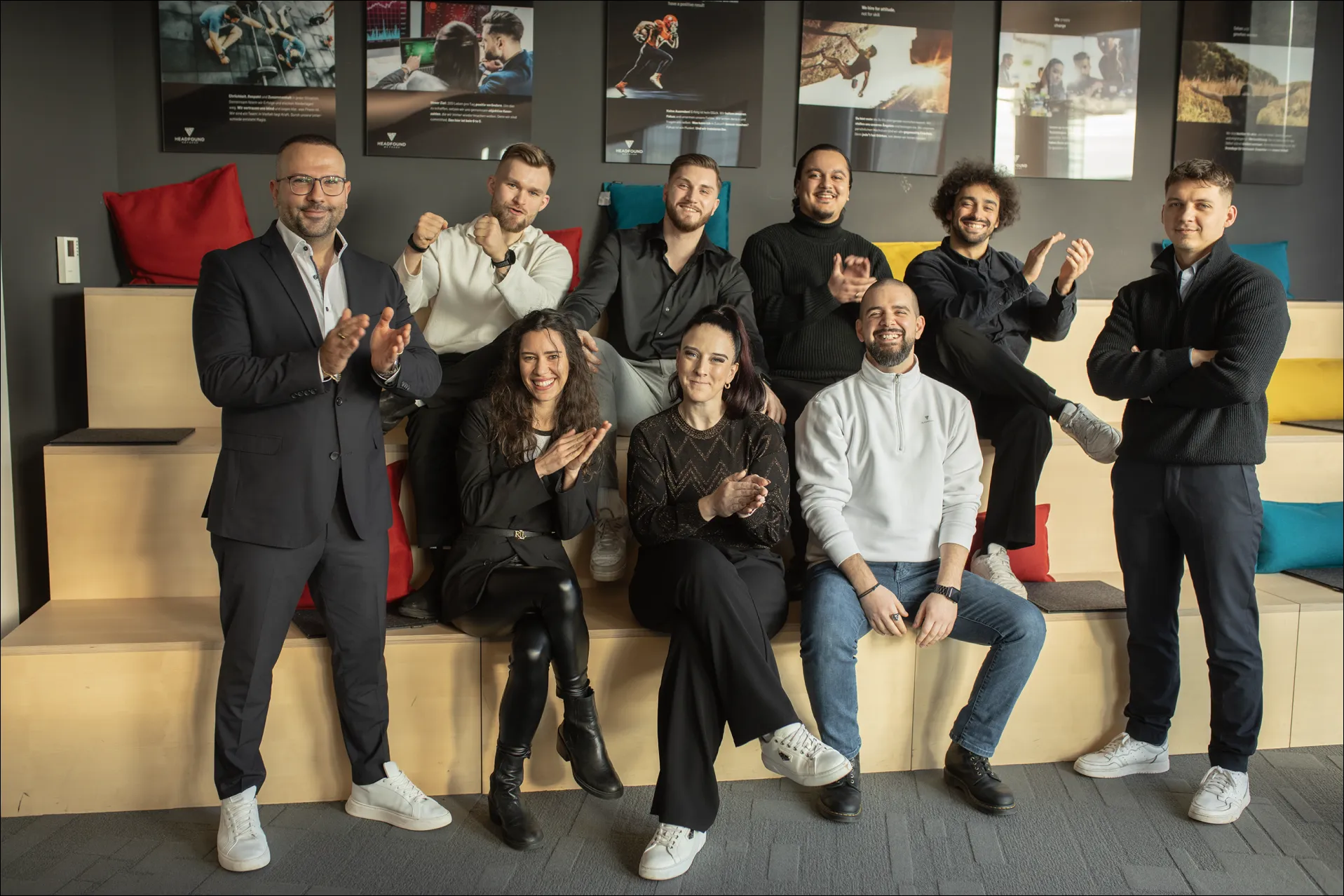 Ein gemischtes Team von neun Personen sitzt und steht lachend auf hölzernen Stufen mit bunten Kissen.