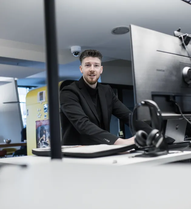 Junger Mann in schwarzem Sakko sitzt an einem Schreibtisch mit Computer in modernem Büro.