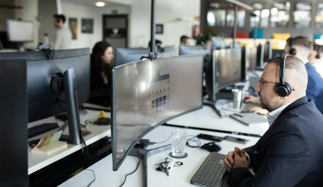 Mann mit Kopfhörern arbeitet an gekrümmtem Bildschirm in einem modernen Büro mit mehreren Arbeitsplätzen.