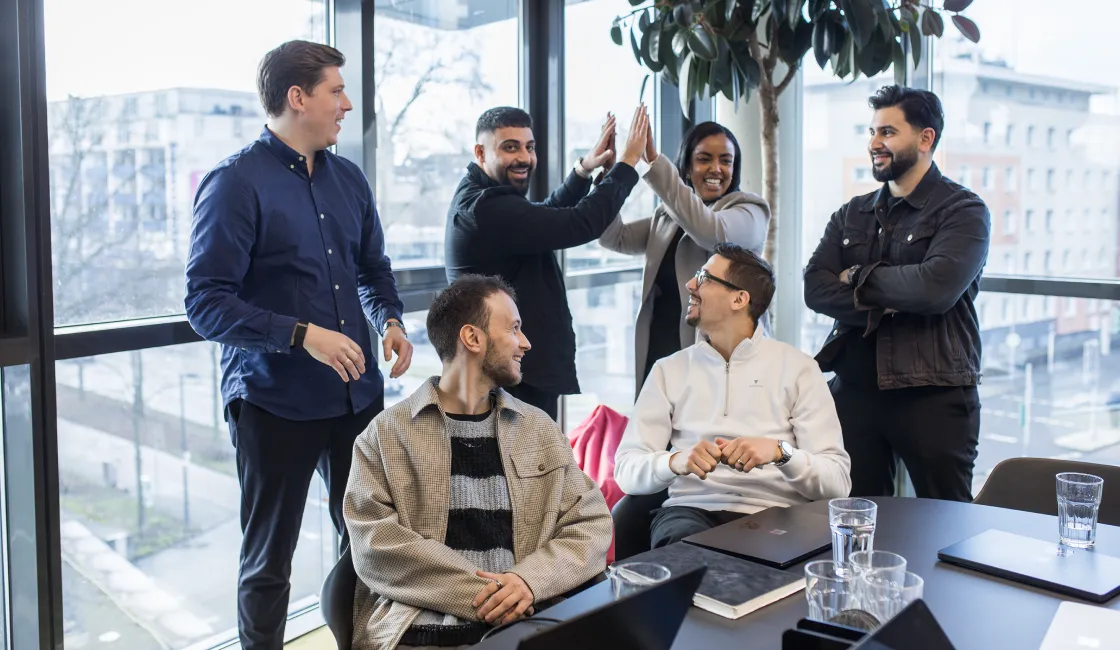 Sechs Kollegen in einem Büro, zwei geben sich high-five, andere lächeln und unterhalten sich.