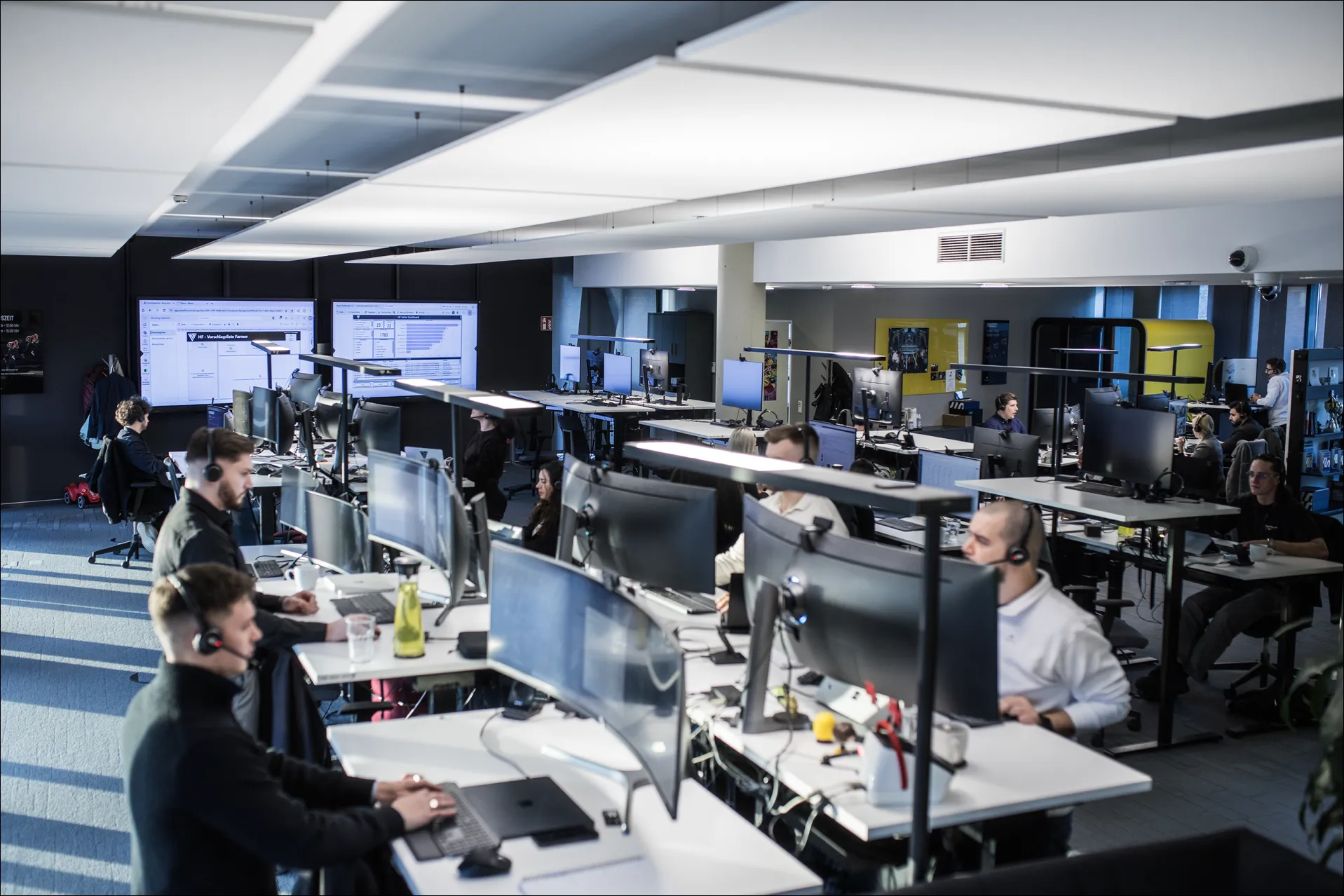 Büro mit Mitarbeitern an Computerarbeitsplätzen, einige mit Headsets, und großen Bildschirmen an der Wand.