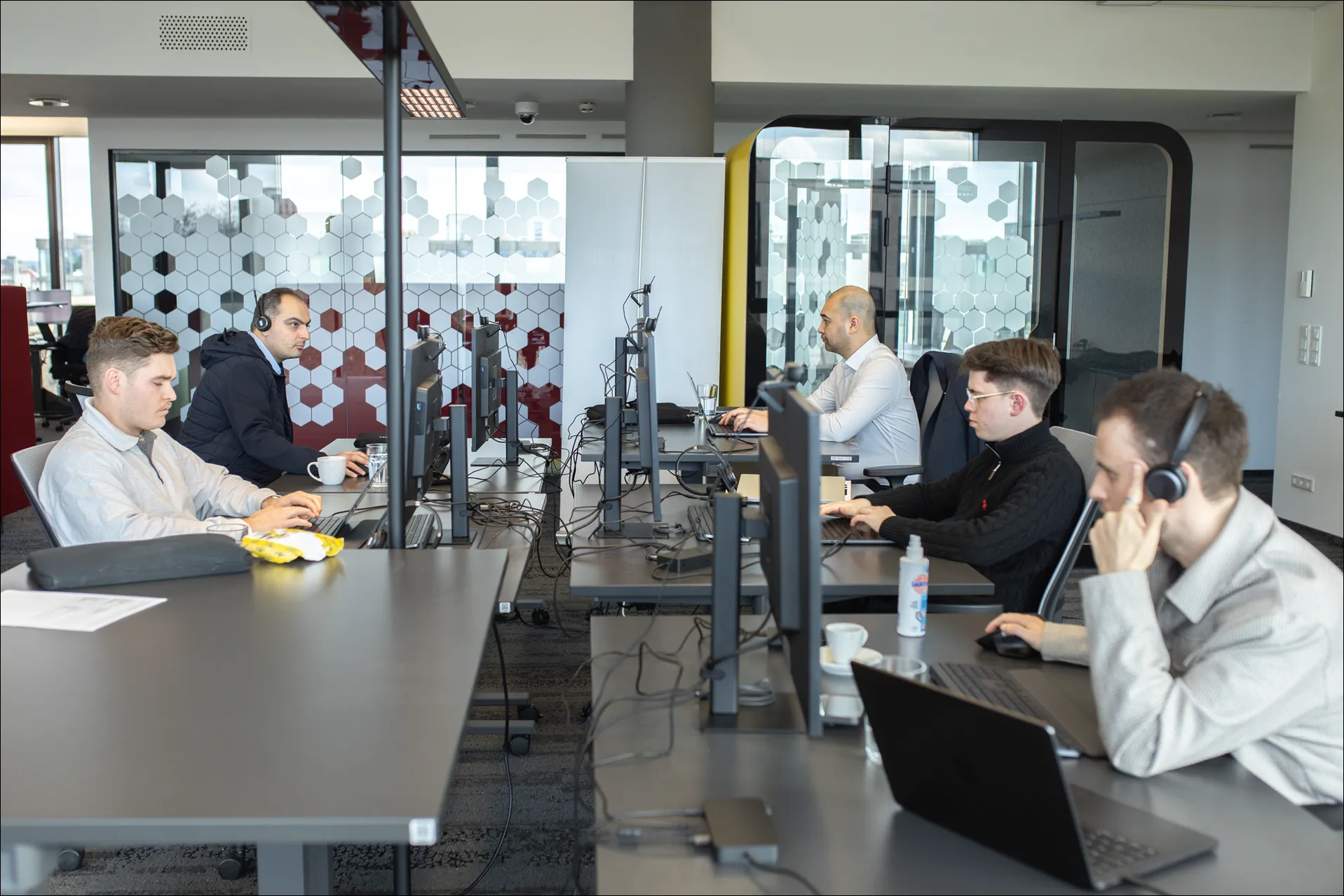 Fünf Männer arbeiten konzentriert an Computern in einem modernen Büro mit gläsernen Trennwänden.