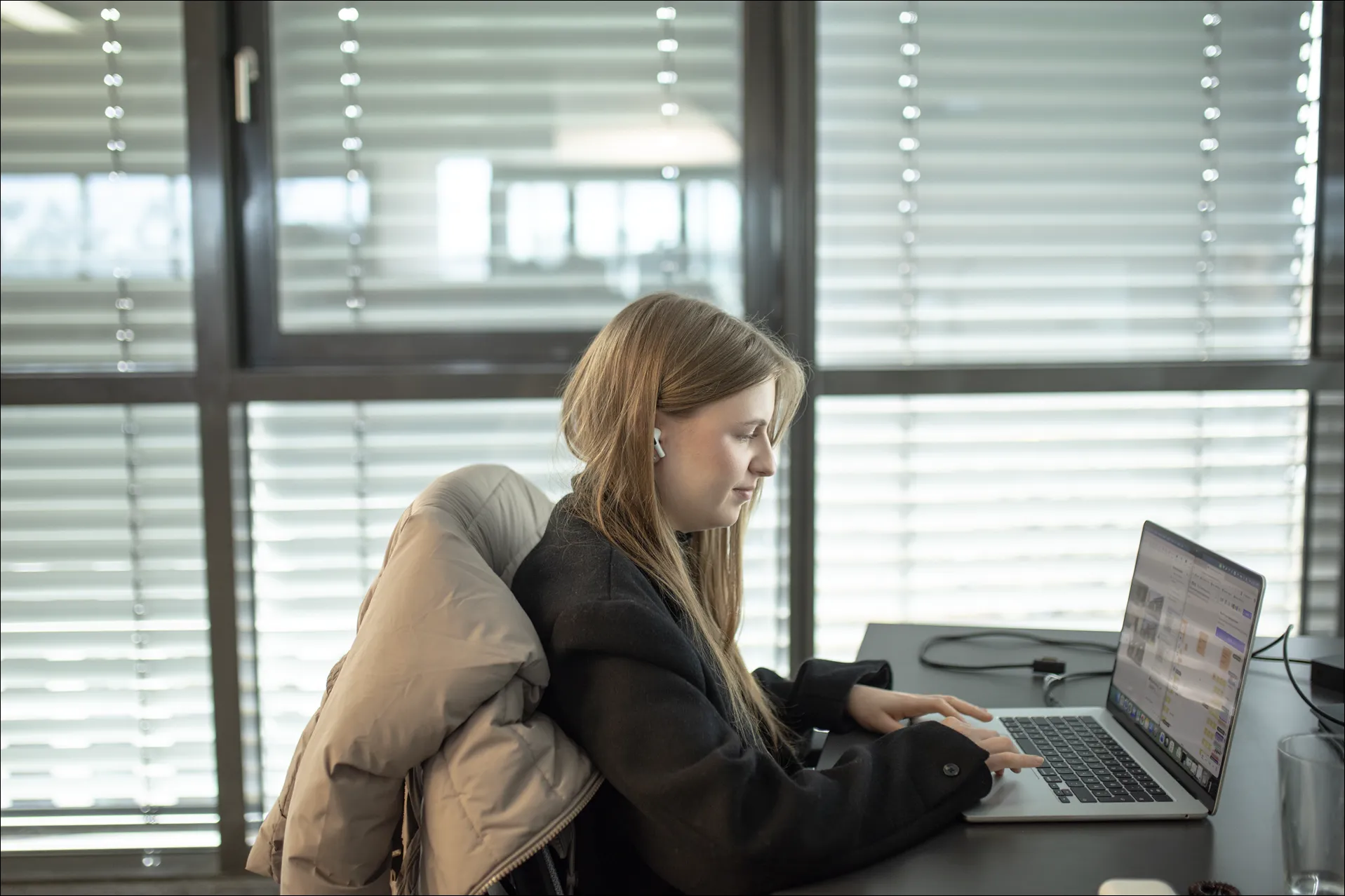 Frau mit langem Haar trägt Ohrhörer und arbeitet an Laptop in einem hellen Büro.