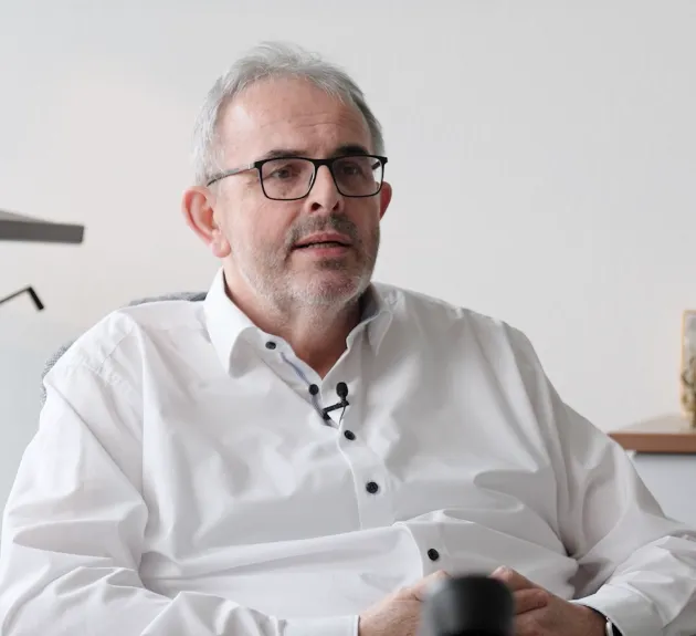 Älterer Mann mit Brille und weißem Hemd, der in einem Stuhl sitzt und spricht.