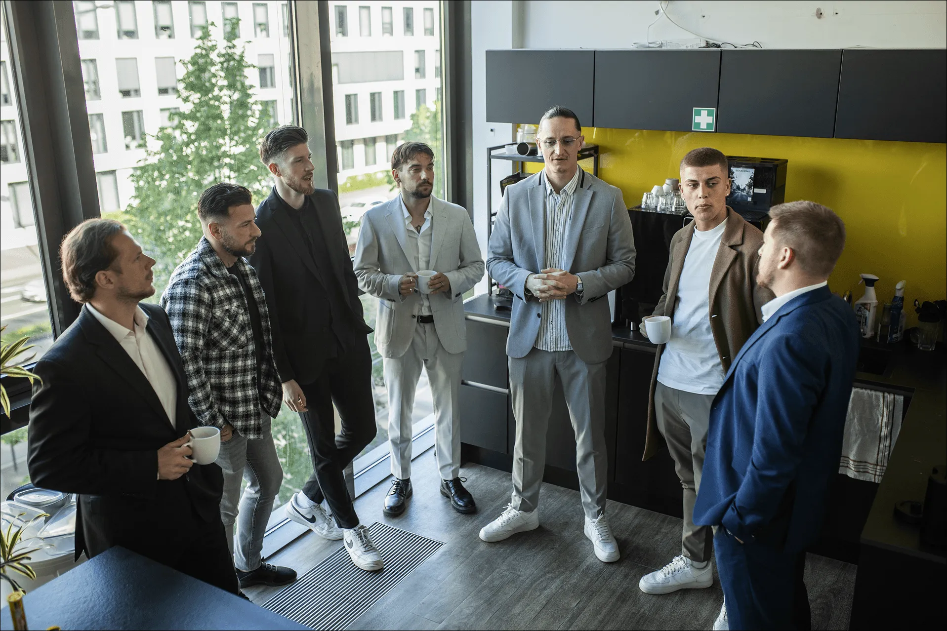 Sieben Männer stehen in einem modernen Büro und unterhalten sich bei Kaffee.
