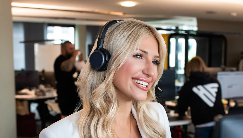 Frau mit Headset und Smartwatch blickt aus Fenster, stützt sich auf Tisch, inneres Büro.