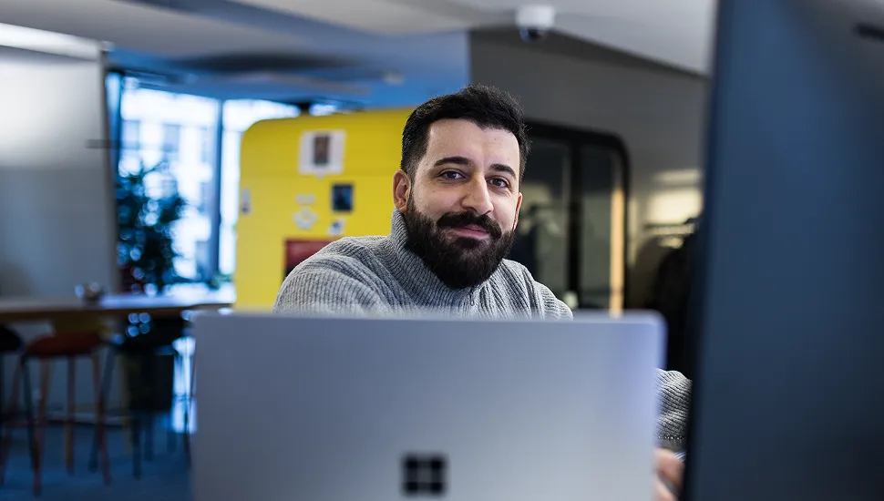 Mann mit Bart und grauem Pullover lächelt am Laptop in einem modernen Büro.
