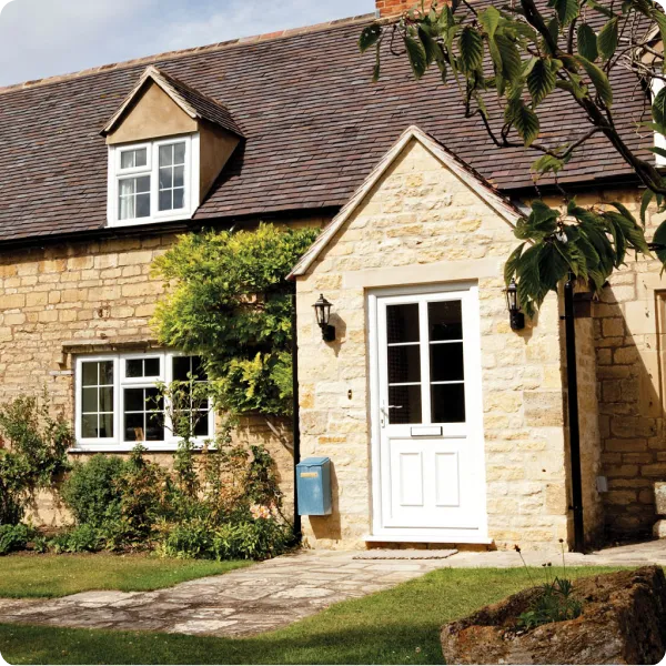 Stone cottage with a white door featuring glass panels, white-framed windows, and greenery along the front yard.