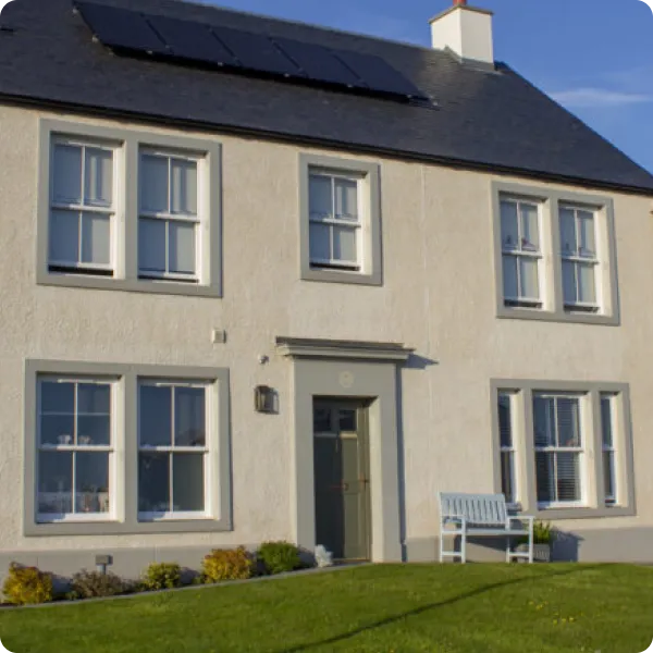 Two-story beige house with white-framed double-hung windows, a front door, solar panels on the roof, and a blue bench on the lawn.
