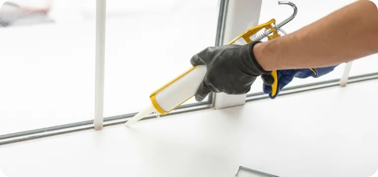 Person wearing a black glove applying white caulk along a window frame using a caulking gun.