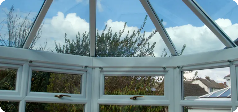 View through a white-framed glass conservatory roof and windows showing leafy bushes and partly cloudy blue sky outside.