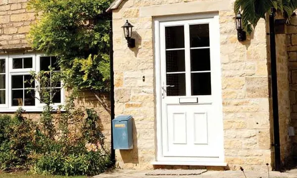 White front door with glass panes set in a beige stone house, flanked by two black exterior wall lights and a blue mailbox, with greenery nearby.
