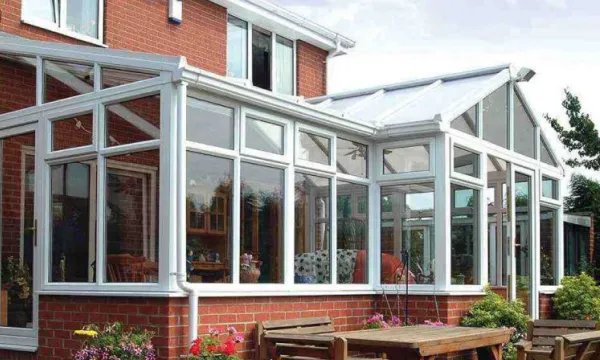 White-framed glass conservatory attached to a red brick house with outdoor wooden table and benches nearby.