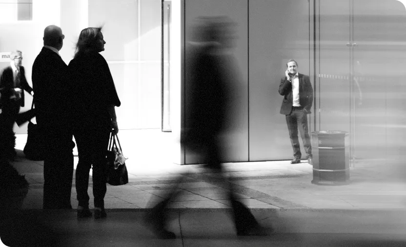 Black and white photo of people walking and standing in an urban setting, featuring a man talking on the phone near a building and blurred figures in motion.
