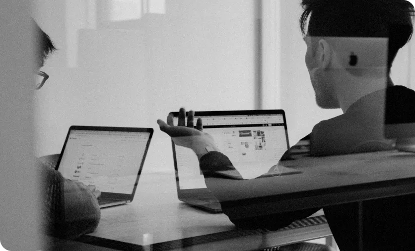 Two people sitting at a table working on laptops, one gesturing toward the screen during a discussion.