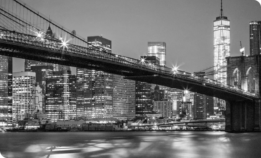 Black and white photo of the Brooklyn Bridge spanning over the East River with the illuminated Manhattan skyline in the background at dusk.
