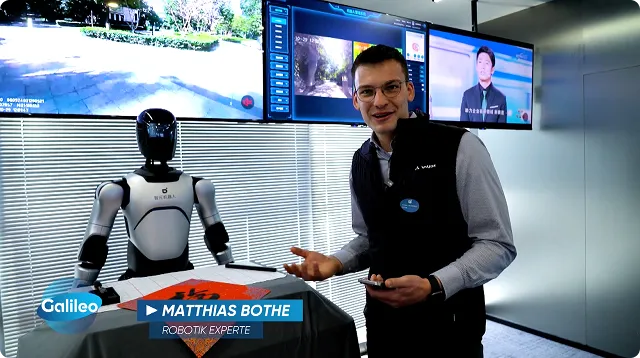 Mann mit Brille und namensschild präsentiert einen weißen humanoiden Roboter neben einem Tisch mit einem orangen Tuch, im Hintergrund mehrere Monitore.