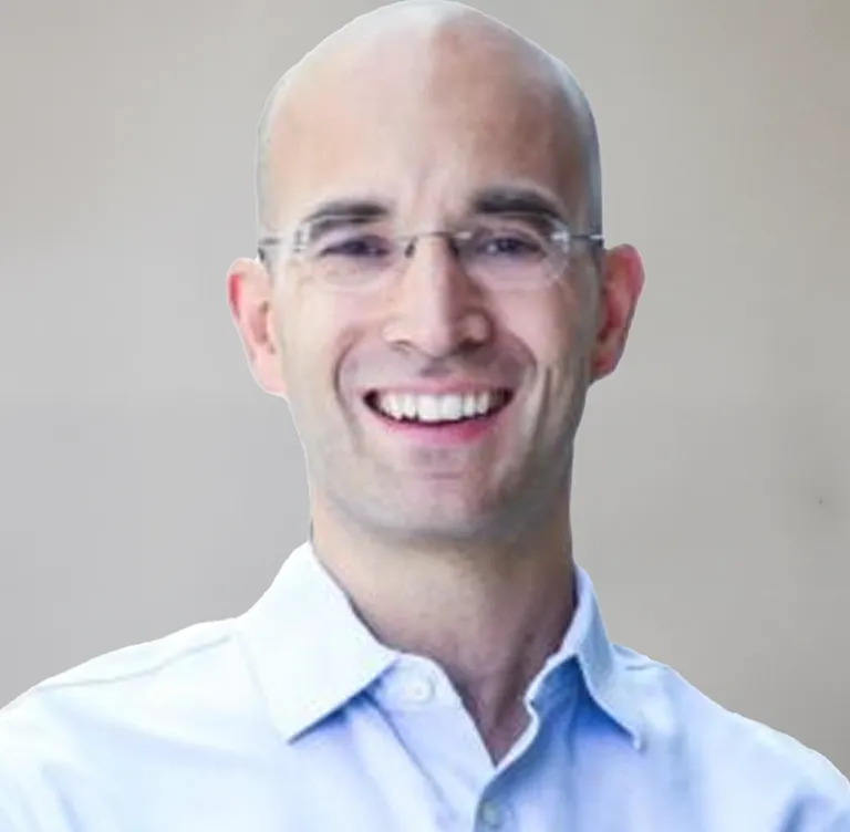 Porträt eines lächelnden, glatzköpfigen Mannes mit Brille und blauem Hemd vor neutralem Hintergrund.