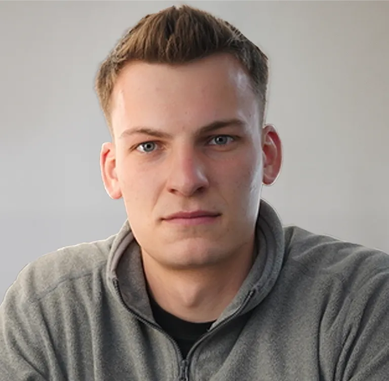 Junger Mann mit kurzen blonden Haaren und grauem Pullover vor unscharfem hellgrauem Hintergrund.