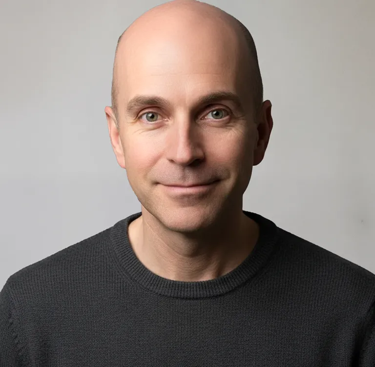 Porträt eines lächelnden, glatzköpfigen Mannes mit grünlichen Augen, der einen schwarzen Pullover trägt.