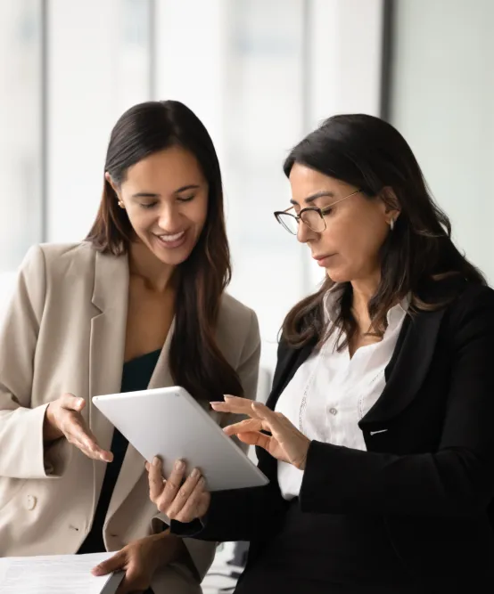Two professional women in blazers looking at a tablet together in a bright office.