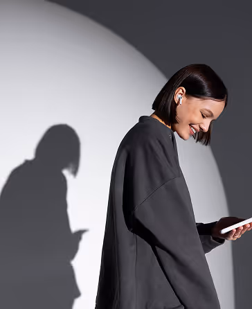Smiling woman with short dark hair wearing wireless earbuds and a dark oversized sweatshirt, looking at her phone with her shadow cast on a gray wall behind her.