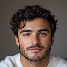 Portrait of a young man with dark curly hair, beard, and mustache, wearing a light-colored top against a neutral background.
