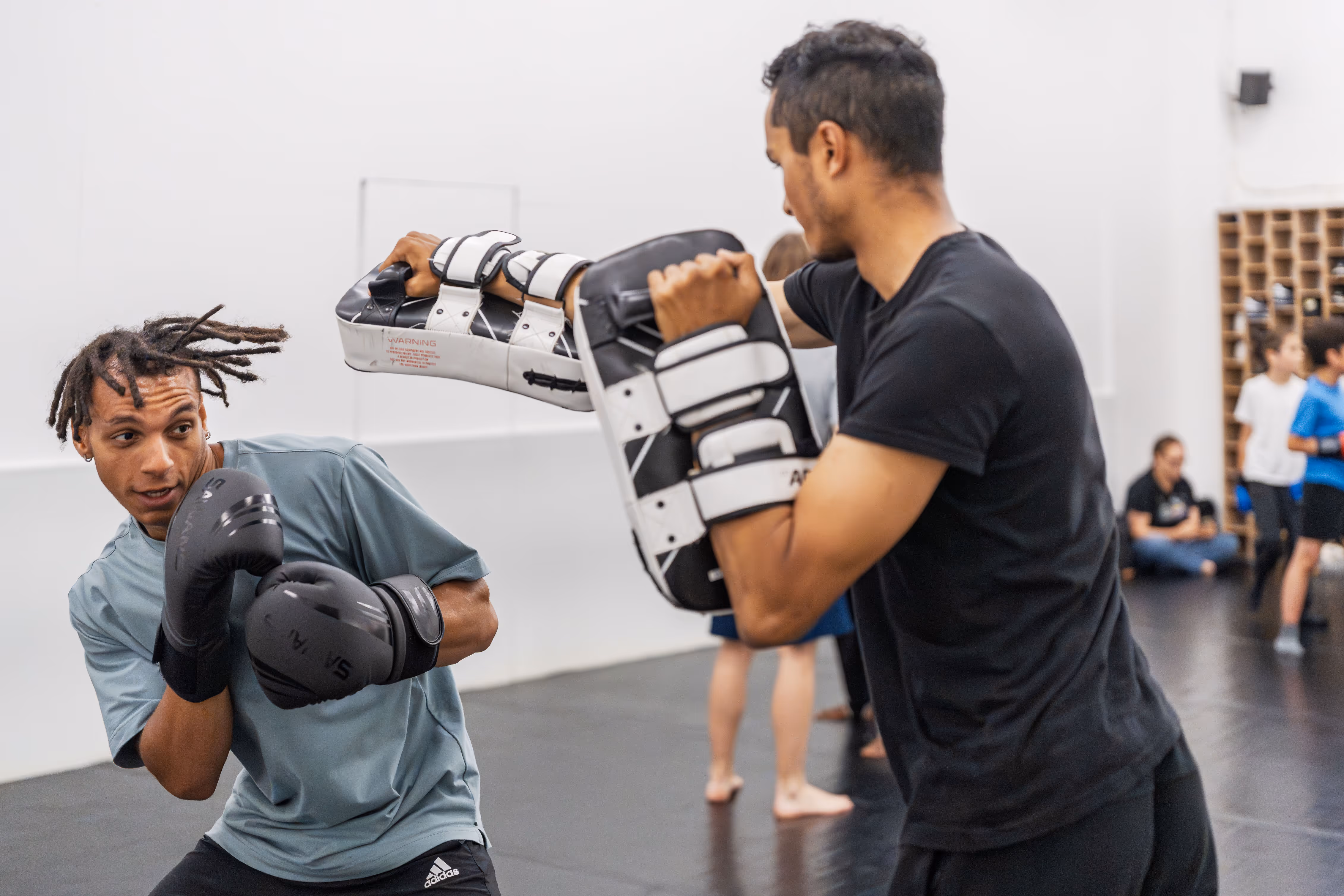 Boxer dodging a punch from a trainer holding black and white focus pads in a gym.