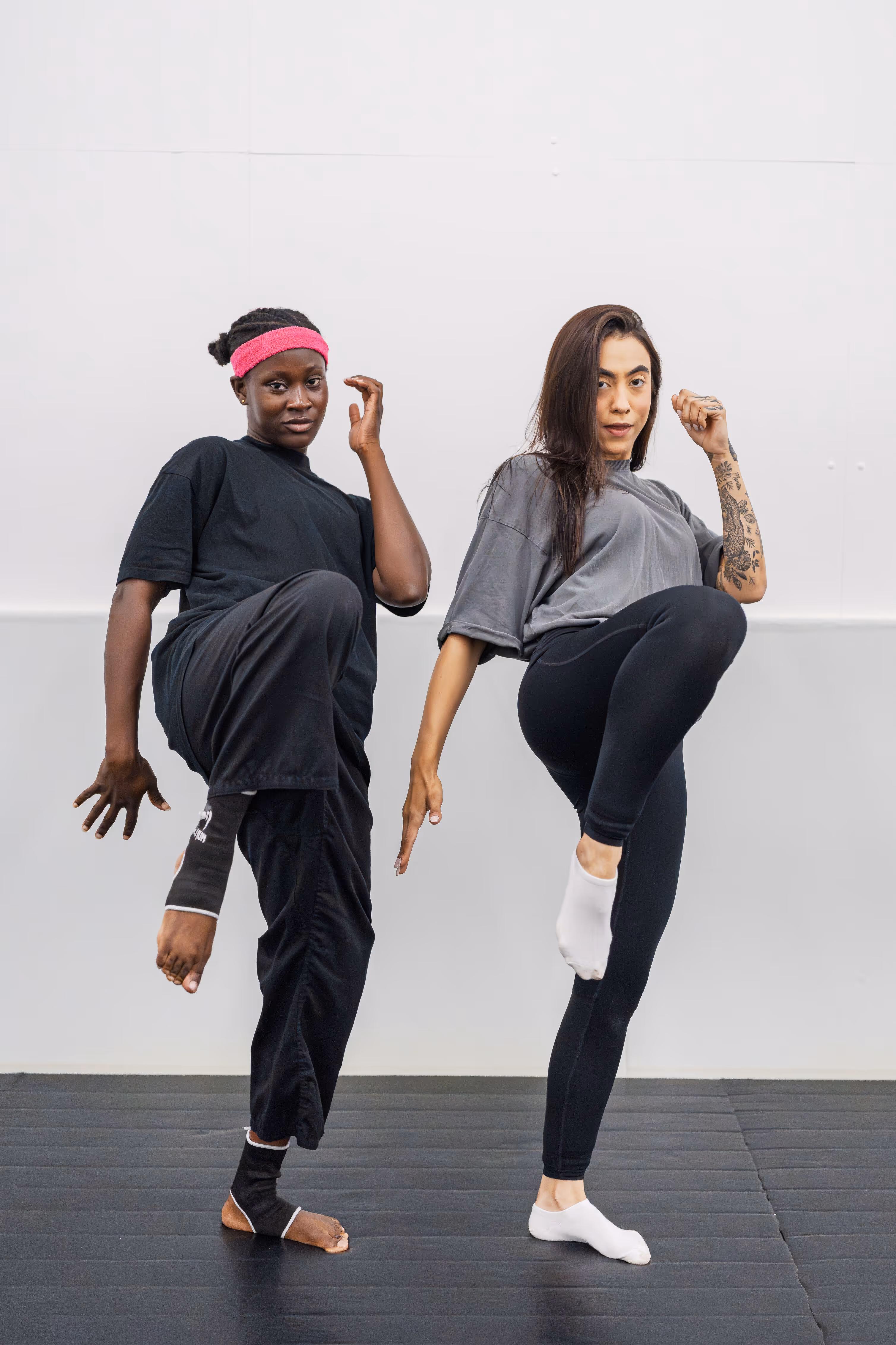 Two women practicing martial arts kicks in a studio with a white wall and black floor mats.