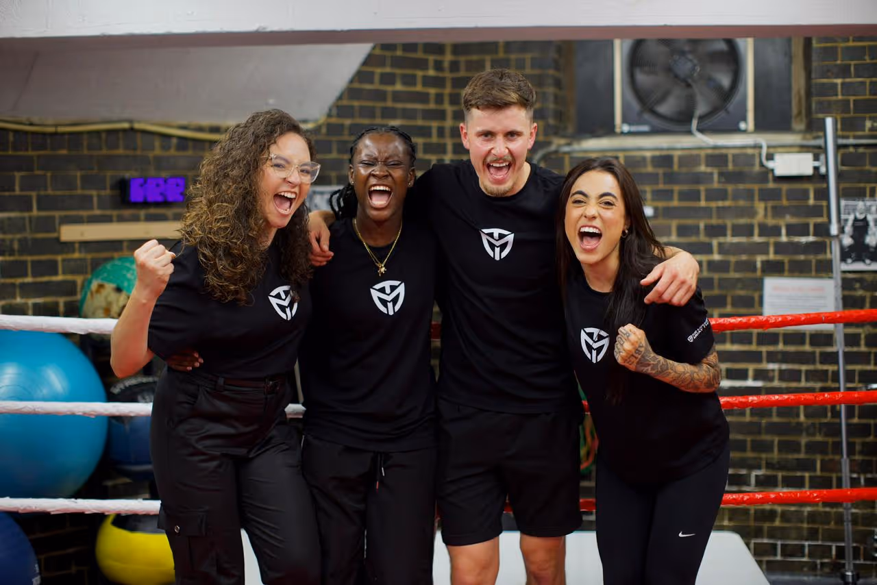 Four excited adults in black sportswear cheering together inside a gym boxing ring.