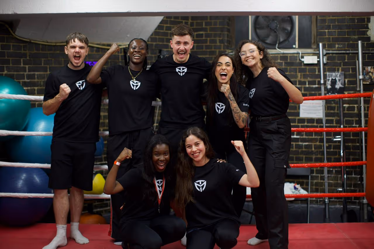 Group of seven diverse people in matching black shirts cheer with fists raised inside a boxing ring.