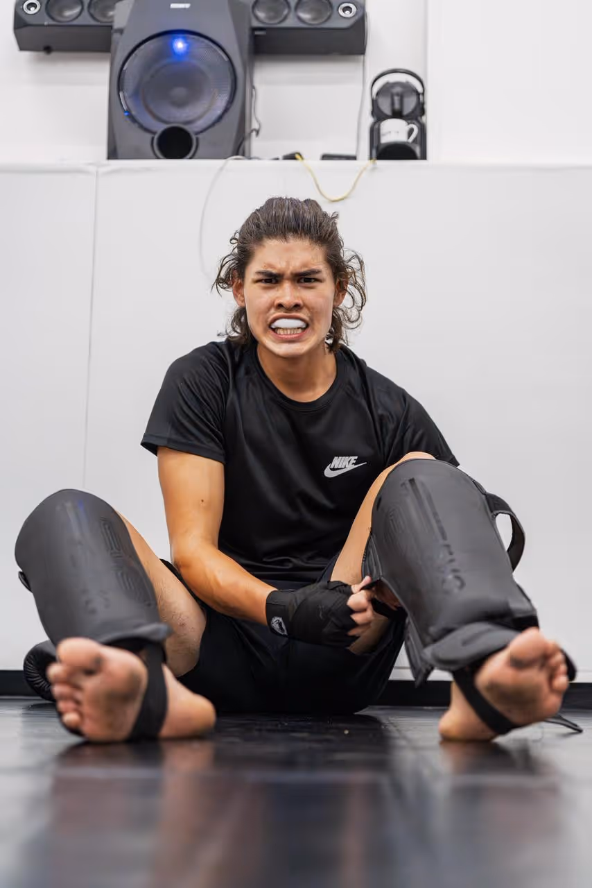 Young man in black Nike shirt sitting on floor wearing black shin guards, making an intense face.