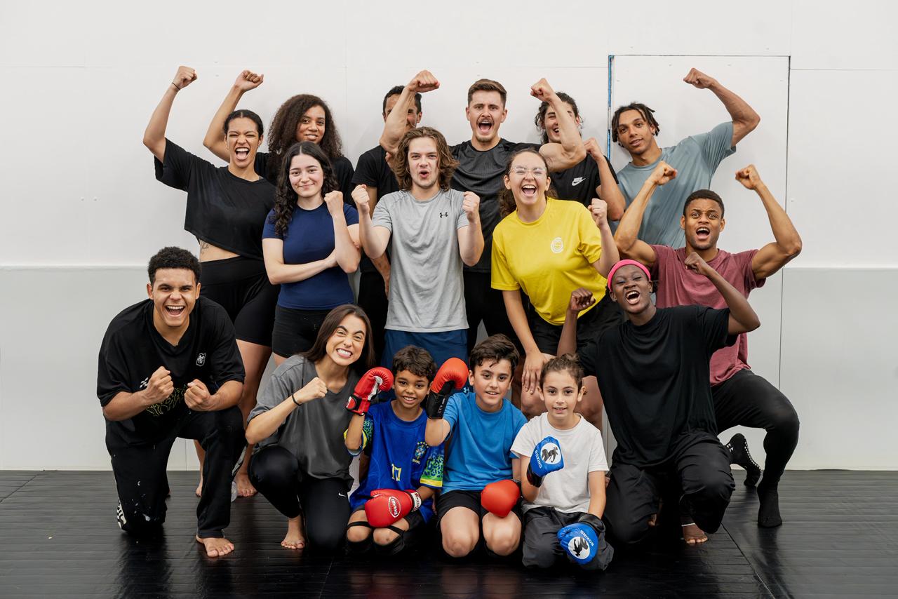 Group of diverse young people and kids posing enthusiastically with fists raised, some wearing boxing gloves.