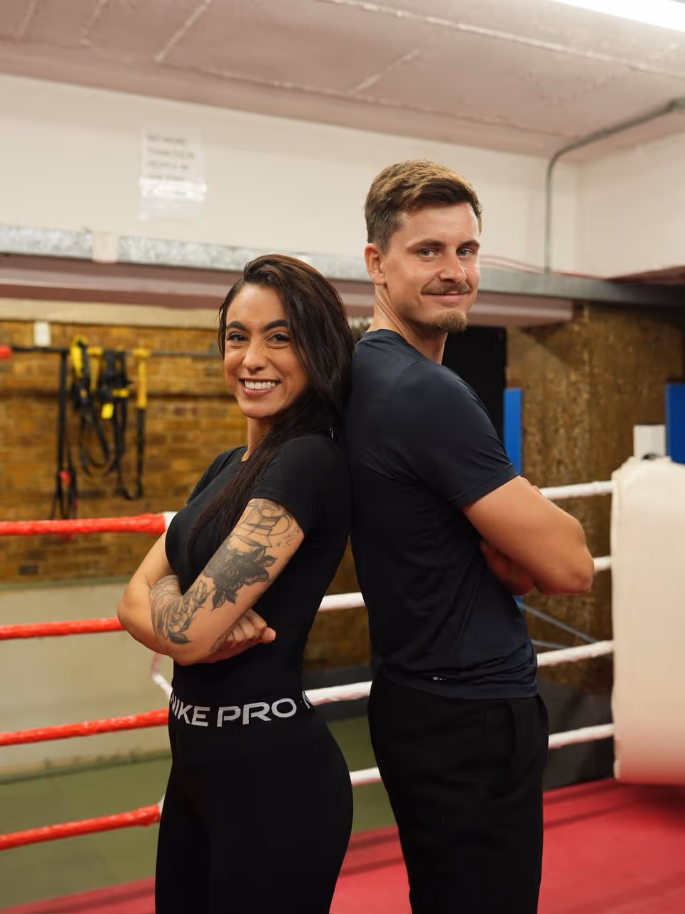 Smiling man and woman stand back to back with arms crossed in a boxing gym ring.