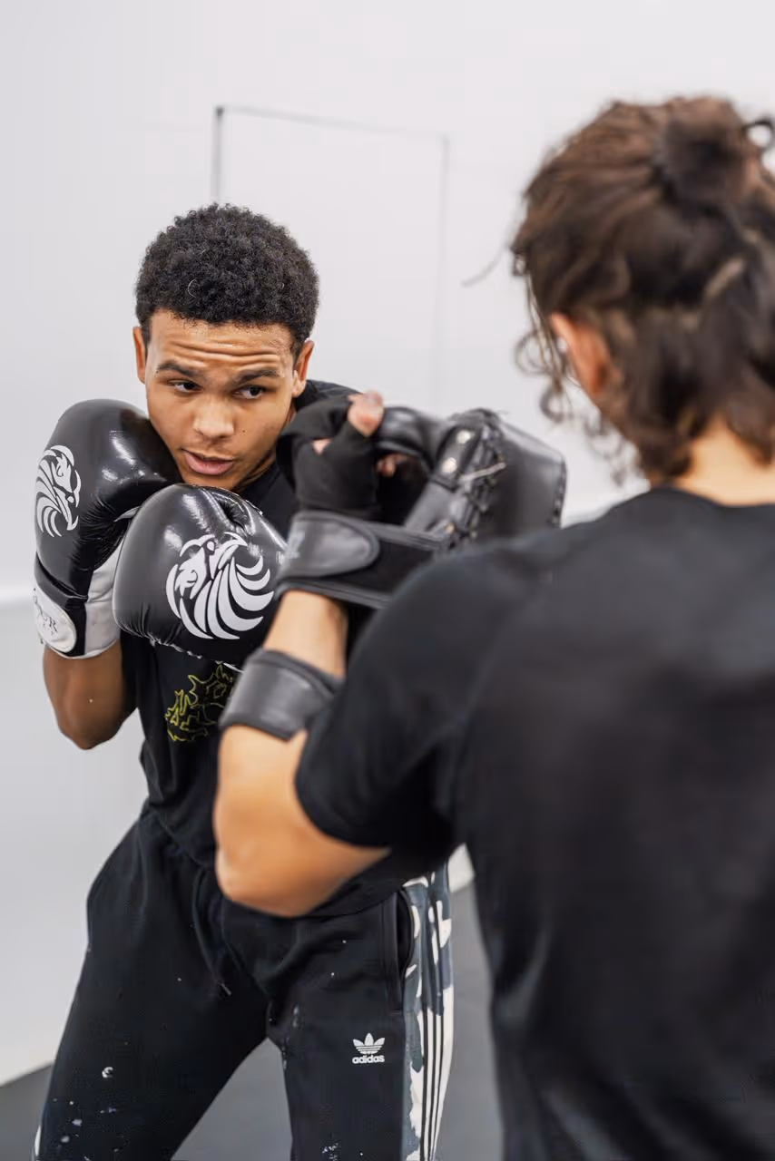 Two men sparring in a boxing gym, one in black gloves preparing to punch a shield.
