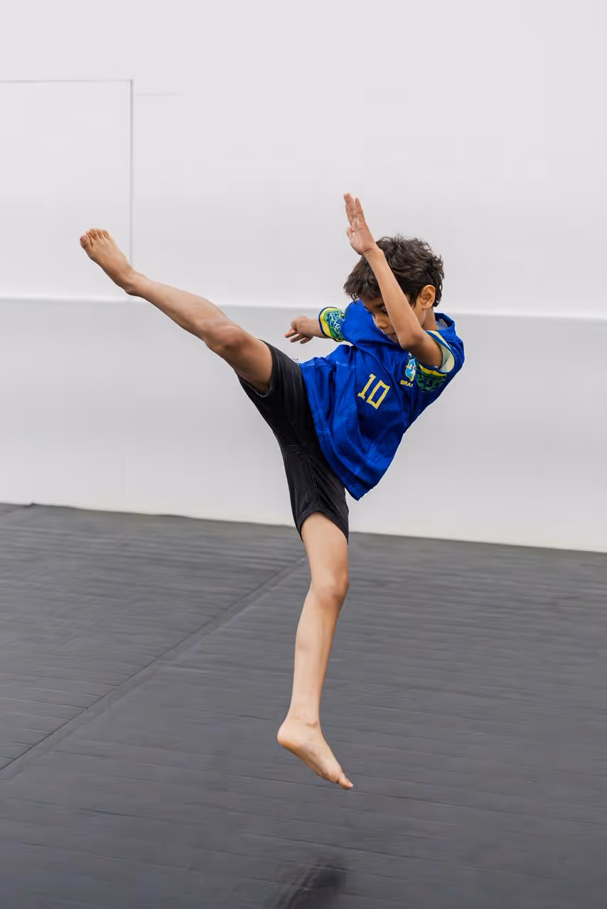 Boy in blue sports jersey doing a high kick in an indoor gym with padded flooring.
