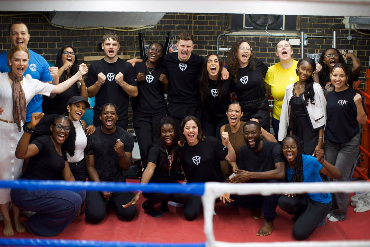 Diverse group of people cheering inside a boxing ring with fists raised and big smiles.