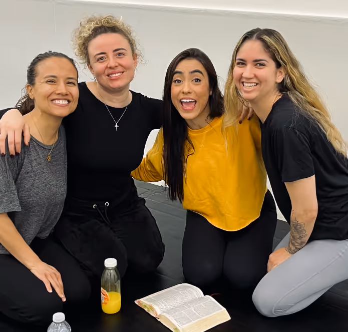Four smiling women sitting close together on a mat with an open book and drinks in front of them.