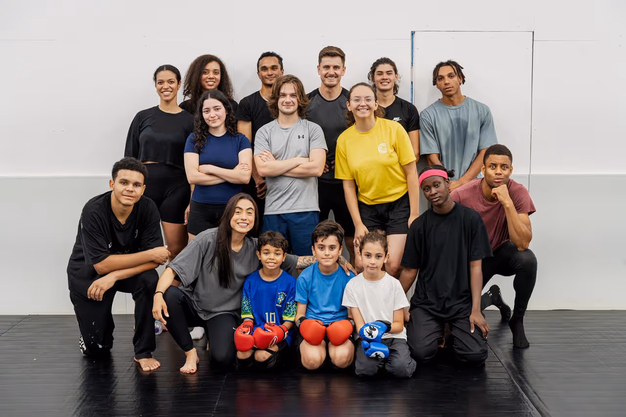 A diverse group of 15 people posing on a black floor against a white wall, including children with boxing gloves.