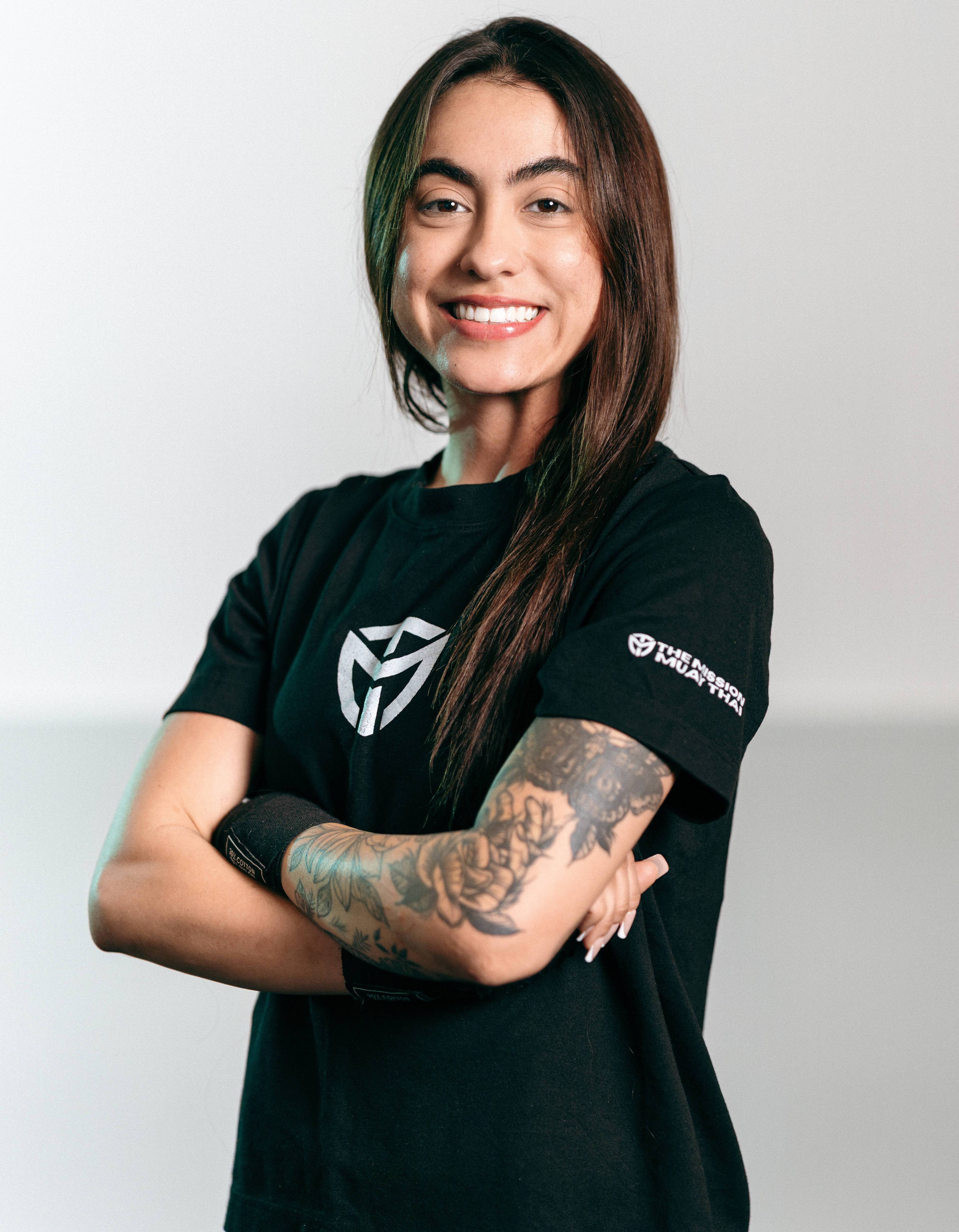 Smiling woman with long hair and floral tattoo on arm wearing a black shirt with Muay Thai logo.