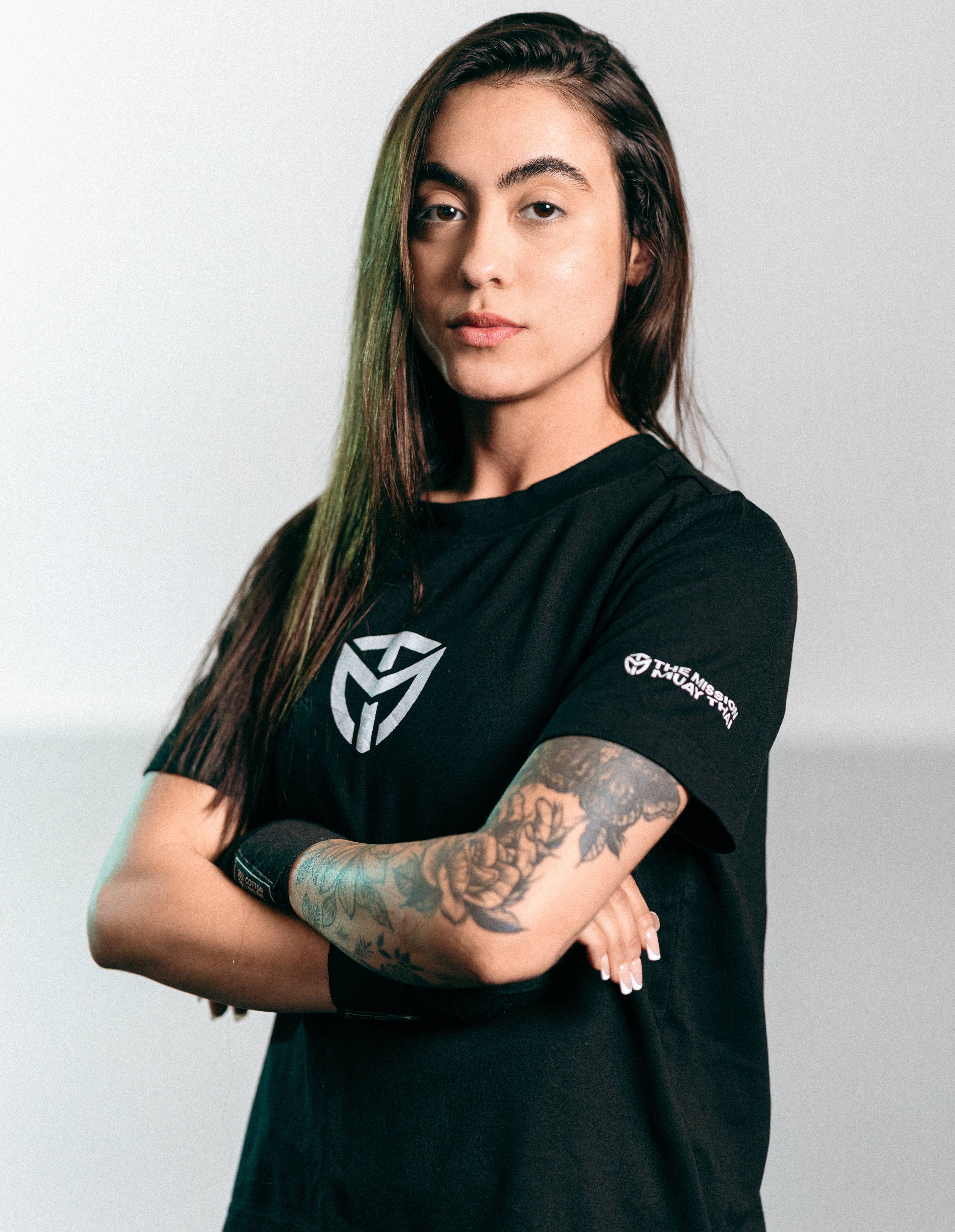 Confident woman with long hair and arm tattoos wearing a black shirt with crossed arms.
