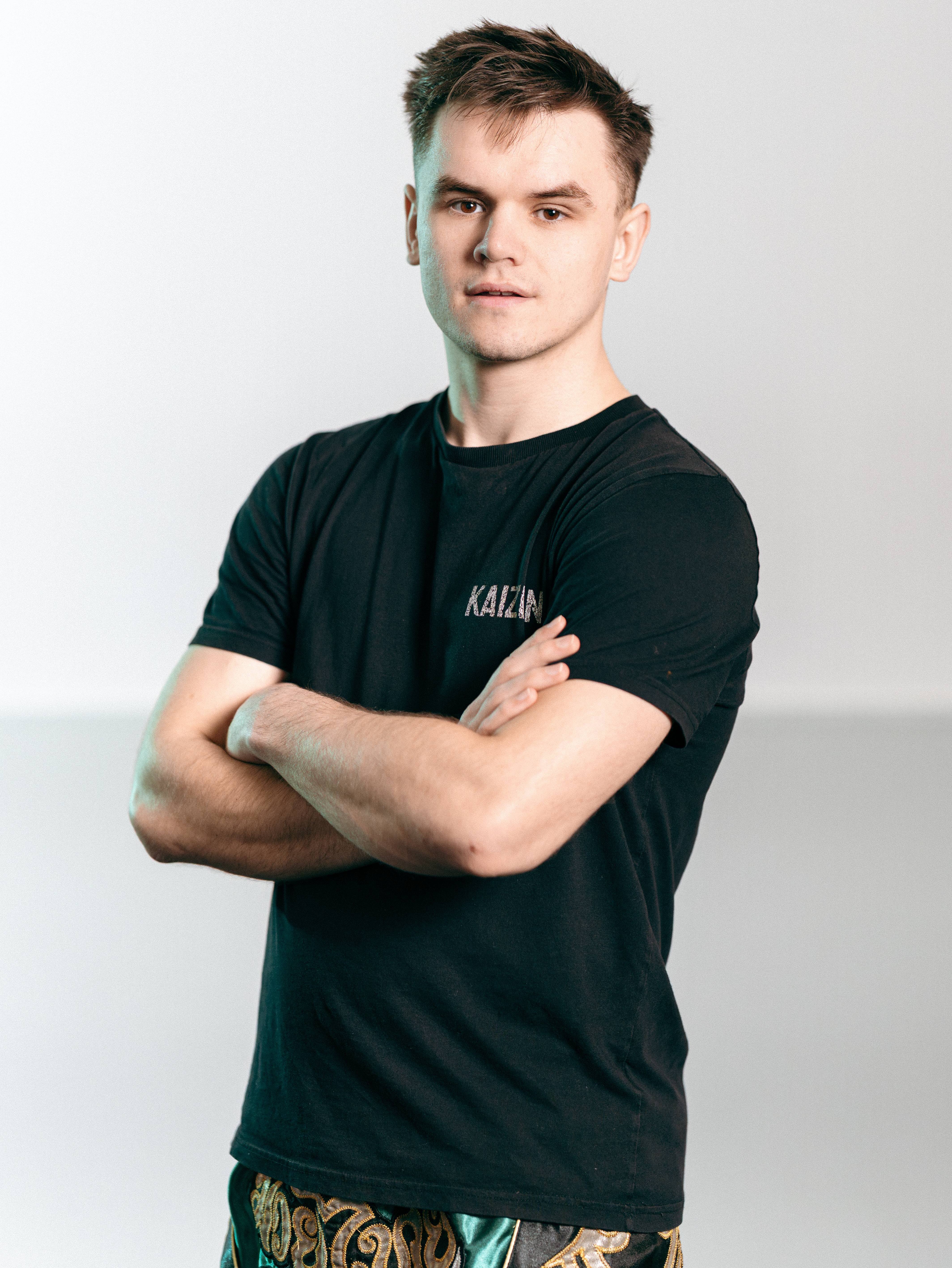 Young man with short hair wearing a black shirt and patterned shorts, arms crossed, looking at camera.