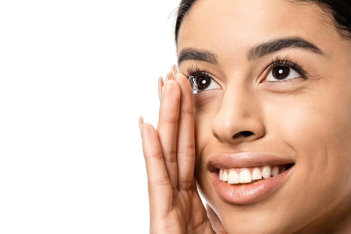 Close-up of a smiling woman holding a contact lens near her eye, preparing to insert it.